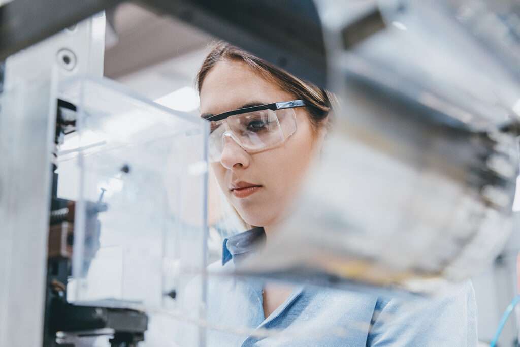 Konzentrierte Laborantin mit Schutzbrille inspiziert Maschinen und stellt die Qualität sicher.